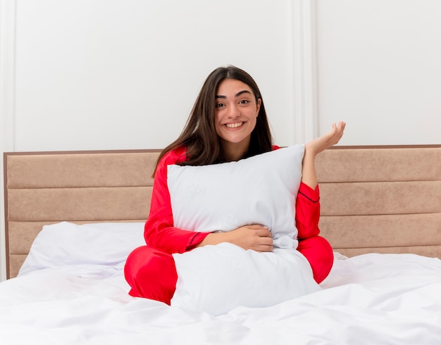 Foto gratuita mujer hermosa joven en pijama rojo sentado en la cama con almohada feliz y positivo sonriendo alegremente en el interior del dormitorio