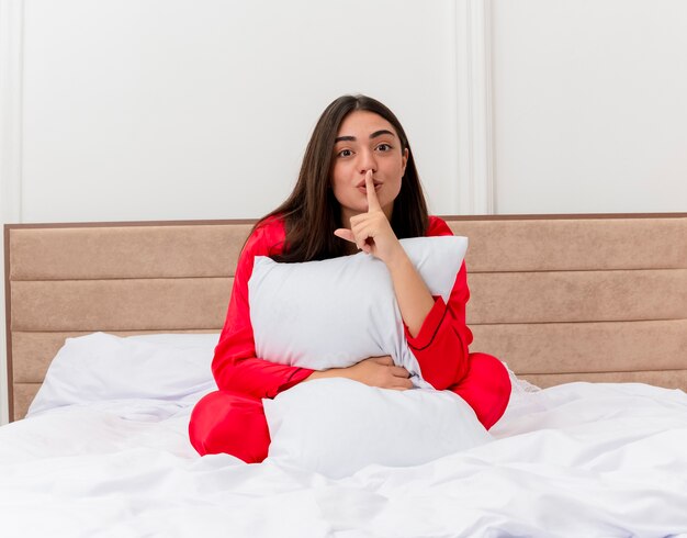 Mujer hermosa joven en pijama rojo sentado en la cama con almohada haciendo gesto de silencio con el dedo en los labios en el interior del dormitorio