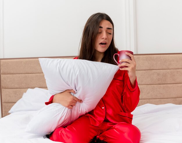 Mujer hermosa joven en pijama rojo sentado en la cama con almohada sosteniendo una taza con aspecto cansado sensación fatiga matutina en el interior del dormitorio sobre fondo claro