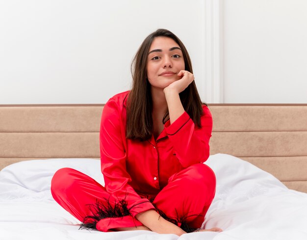 Mujer hermosa joven en pijama rojo sentado en la cama con la cabeza inclinada en la mano mirando a la cámara sonriendo en el interior de la casa sobre fondo claro