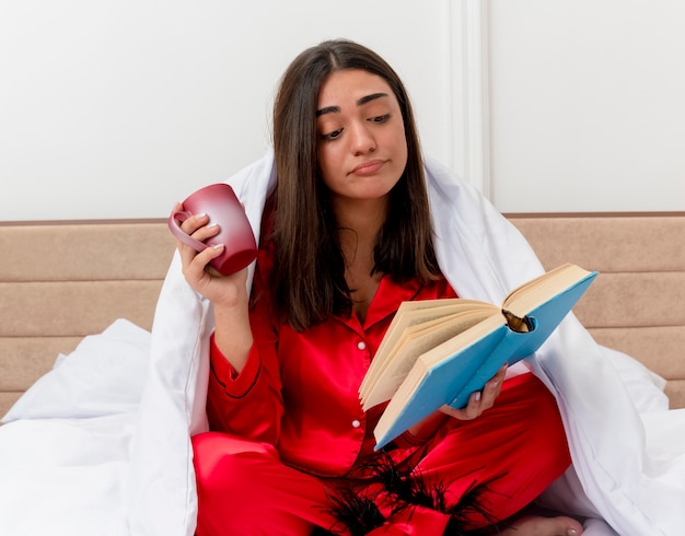 Mujer hermosa joven en pijama rojo sentado en la cama envuelto en una manta con una taza de libro de lectura de café en el interior del dormitorio sobre fondo claro