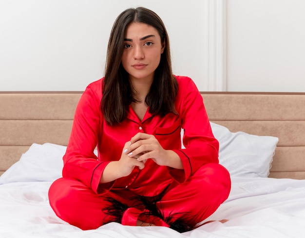 Mujer hermosa joven en pijama rojo sentado en la cama mirando a cámara con cara seria en el interior del dormitorio sobre fondo claro