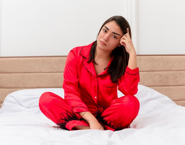 Mujer hermosa joven en pijama rojo sentado en la cama mirando a cámara con cara seria en el interior del dormitorio sobre fondo claro