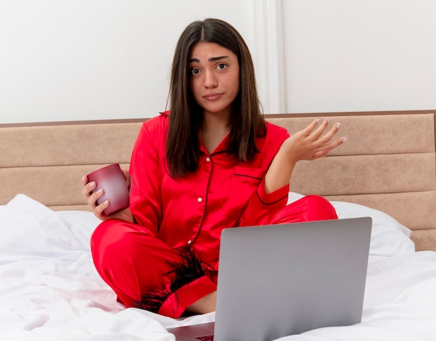 Mujer hermosa joven en pijama rojo sentado en la cama con un portátil y una taza de café mirando a la cámara confundido extendiendo el brazo hacia un lado en el interior del dormitorio sobre fondo claro