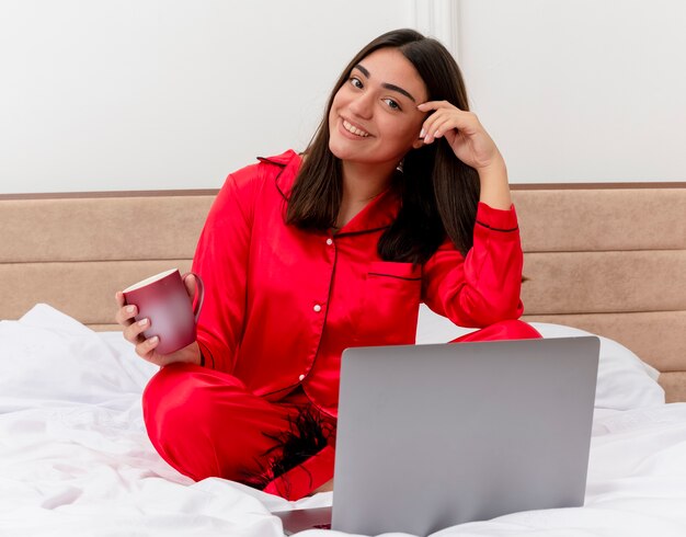 Mujer hermosa joven en pijama rojo sentado en la cama con un portátil y una taza de café mirando a la cámara con una sonrisa en la cara feliz y positiva en el interior del dormitorio sobre fondo claro