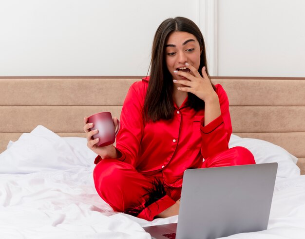 Mujer hermosa joven en pijama rojo sentado en la cama con el portátil y una taza de café mirando la pantalla del portátil sonriendo alegremente en el interior del dormitorio sobre fondo claro