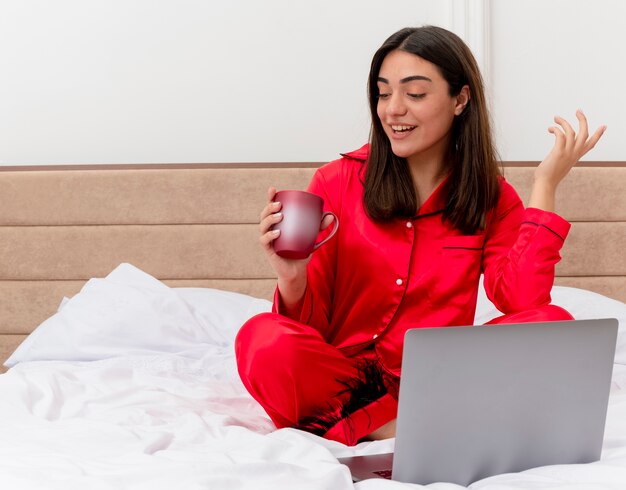 Mujer hermosa joven en pijama rojo sentado en la cama con un portátil y una taza de café mirando la taza sonriendo feliz y positivo en el interior del dormitorio sobre fondo claro