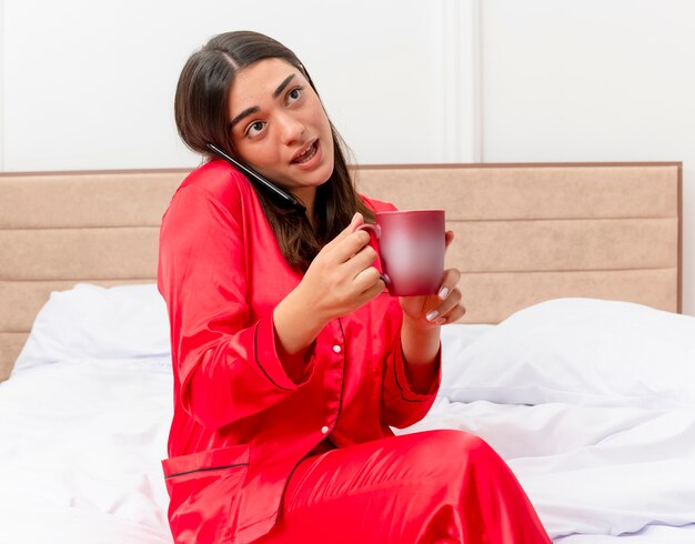 Mujer hermosa joven en pijama rojo sentado en la cama con una taza de café hablando por teléfono móvil en el interior del dormitorio sobre fondo claro