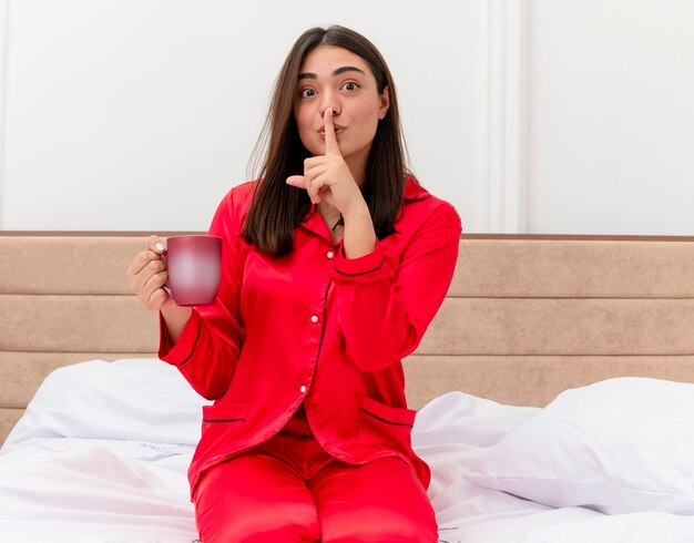 Mujer hermosa joven en pijama rojo sentado en la cama con una taza de café haciendo gesto de silencio con los dedos en los labios en el interior del dormitorio sobre fondo claro