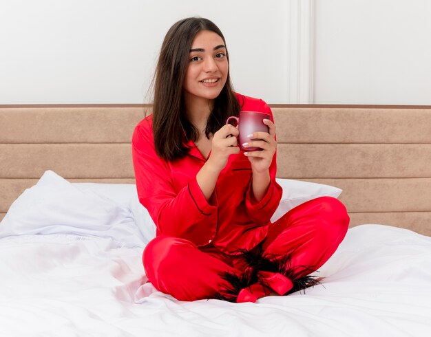 Mujer hermosa joven en pijama rojo sentado en la cama con una taza de café mirando a la cámara con una sonrisa en la cara disfrutando del tiempo en el interior del dormitorio sobre fondo claro