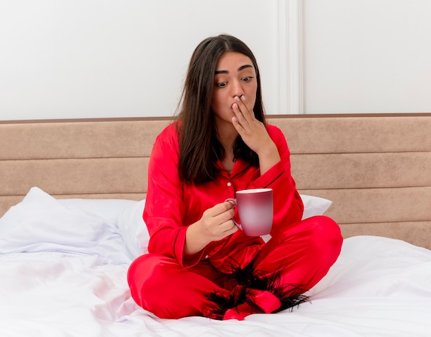 Mujer hermosa joven en pijama rojo sentado en la cama con una taza de café mirando sorprendido cubriendo la boca con la mano en el interior del dormitorio sobre fondo claro