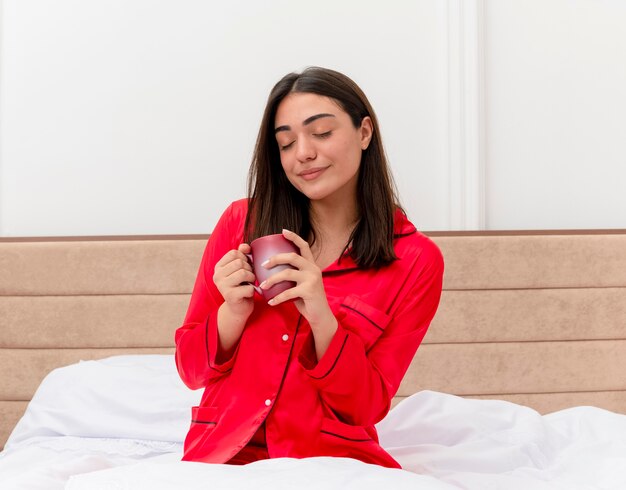 Mujer hermosa joven en pijama rojo sentado en la cama con una taza de café con los ojos cerrados sintiendo emociones positivas en el interior del dormitorio