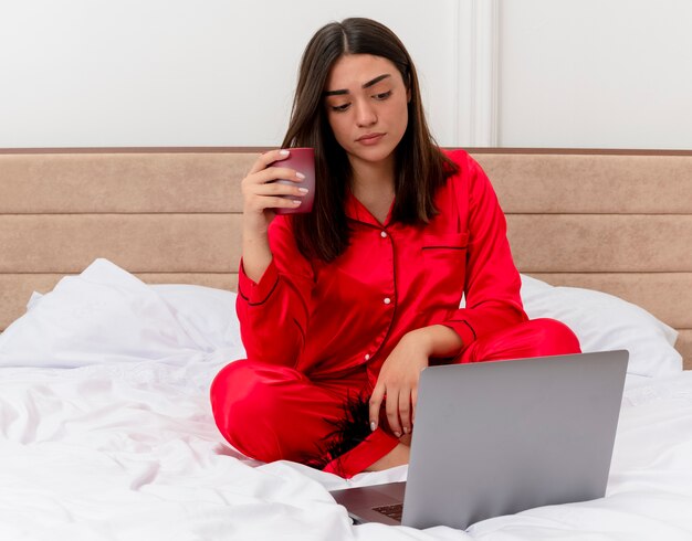 Mujer hermosa joven en pijama rojo sentado en la cama trabajando en la computadora portátil con una taza de café mirando la pantalla con cara seria en el interior del dormitorio sobre fondo claro