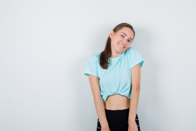 Mujer hermosa joven posando mientras está de pie en camiseta, pantalones y mirando alegre, vista frontal.