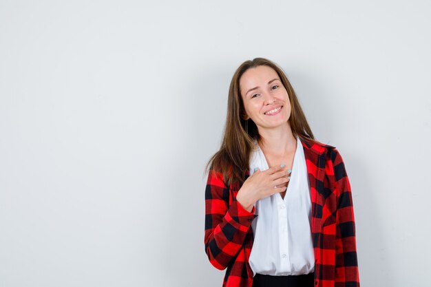 Mujer hermosa joven posando mientras con la mano en el pecho en traje casual y luciendo alegre. vista frontal.