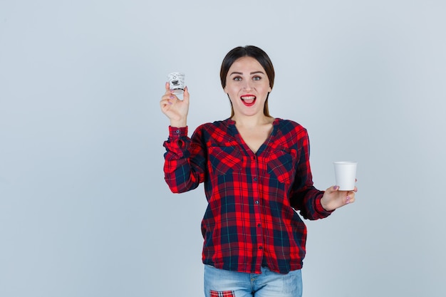 Mujer hermosa joven posando con vasos de plástico en camisa casual, jeans y mirando dichoso, vista frontal.