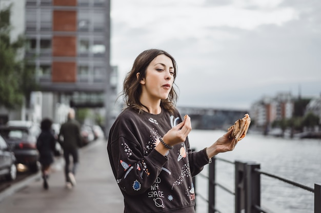 Mujer hermosa joven que come una rebanada de pizza en la calle de la ciudad