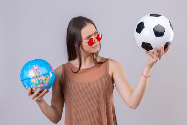 Mujer hermosa joven que lleva gafas de sol rojas que sostienen el balón de fútbol y el globo que mira la bola con expresión sospechosa sobre la pared blanca