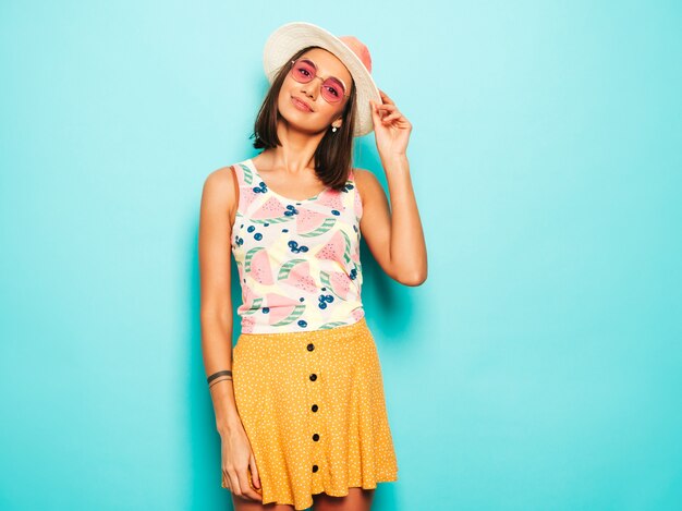 Mujer hermosa joven que mira la cámara en sombrero. Chica de moda en verano casual camiseta blanca y falda amarilla en gafas de sol redondas. La hembra positiva muestra emociones faciales. Modelo divertido aislado en azul