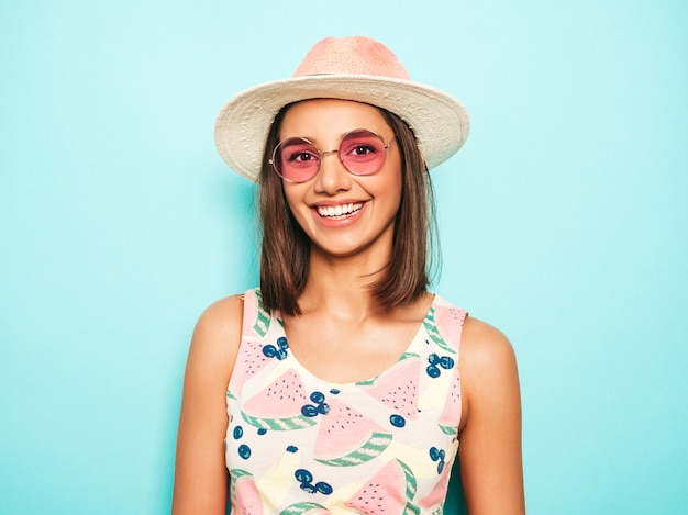 Foto gratuita mujer hermosa joven que mira la cámara en sombrero. chica de moda en verano casual camiseta blanca y falda amarilla en gafas de sol redondas. la hembra positiva muestra emociones faciales. modelo divertido aislado en azul