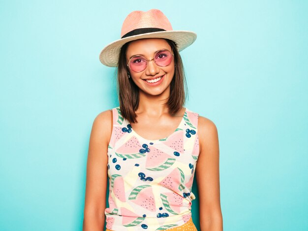 Mujer hermosa joven que mira la cámara en sombrero. Chica de moda en verano casual camiseta blanca y falda amarilla en gafas de sol redondas. La hembra positiva muestra emociones faciales. Modelo divertido aislado en azul