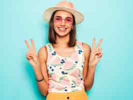 Foto gratuita mujer hermosa joven que mira la cámara en sombrero. chica de moda en verano casual camiseta blanca y falda amarilla en gafas de sol redondas. la hembra positiva muestra emociones faciales. muestra signo de paz