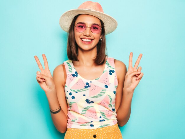 Mujer hermosa joven que mira la cámara en sombrero. Chica de moda en verano casual camiseta blanca y falda amarilla en gafas de sol redondas. La hembra positiva muestra emociones faciales. Muestra signo de paz