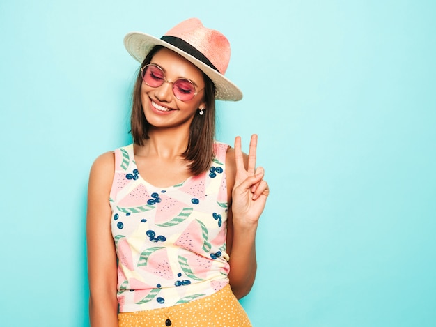 Mujer hermosa joven que mira la cámara en sombrero. Chica de moda en verano casual camiseta blanca y falda amarilla en gafas de sol redondas. La hembra positiva muestra emociones faciales.