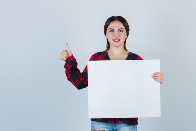 Mujer hermosa joven que muestra el pulgar hacia arriba mientras sostiene el tablero en blanco en camisa casual, jeans y mirando alegre, vista frontal.