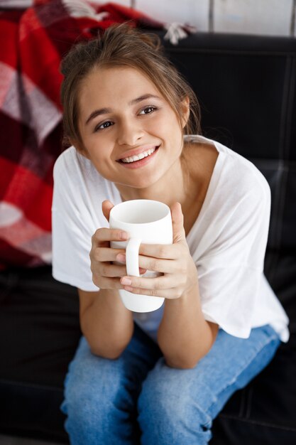 Mujer hermosa joven que sonríe, sosteniendo la taza, sentándose en el sofá.