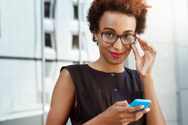 Mujer hermosa joven que sonríe sosteniendo el teléfono caminando por la ciudad