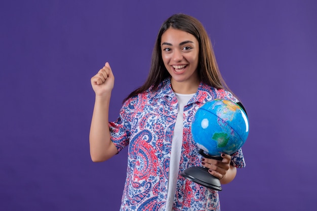 Mujer hermosa joven que sostiene el globo que levanta el puño después de la victoria con la cara feliz que sonríe alegremente, concepto del viaje sobre la pared púrpura