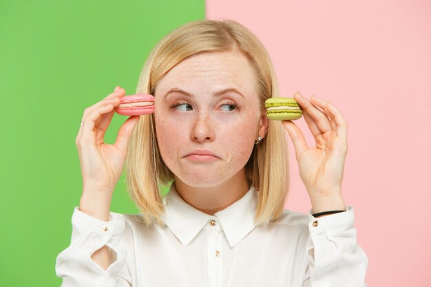 Mujer hermosa joven que sostiene los pasteles de los macarrones en sus manos, sobre coloreado de moda en el estudio.