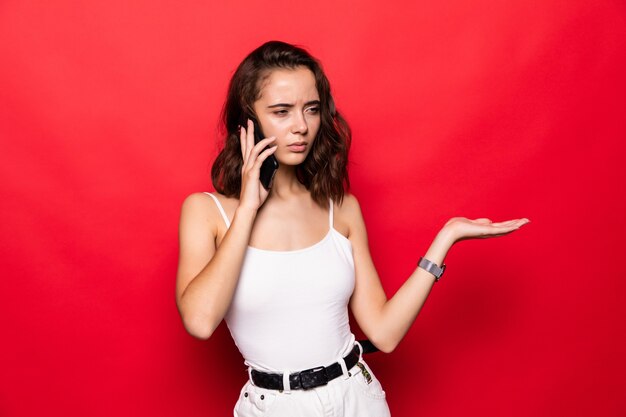 Mujer hermosa joven que tiene conversación hablando por el teléfono inteligente con la mano abierta aislada en la pared roja