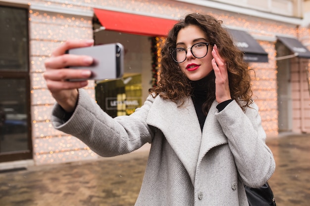 Mujer hermosa joven que toma la foto de Seflie usando el teléfono inteligente, estilo de la ciudad de la calle del otoño, abrigo cálido, gafas, feliz, sonriente, sosteniendo el teléfono en la mano, cabello rizado