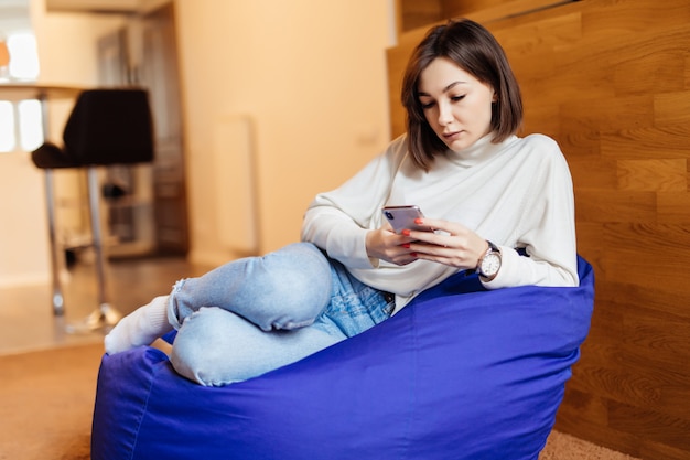 Mujer hermosa joven que usa su teléfono mientras está sentado en el bolso de la silla