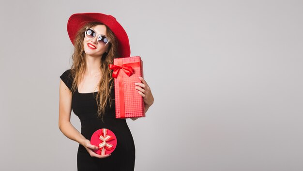 Mujer hermosa joven con regalos, vestido negro, sombrero rojo, gafas de sol, feliz, sonriente, sexy, elegante, cajas de regalo, celebrando, positivo