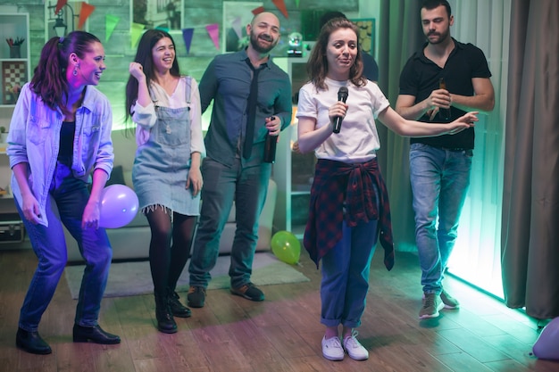 Mujer hermosa joven rodeada de sus amigos haciendo karaoke en la fiesta.