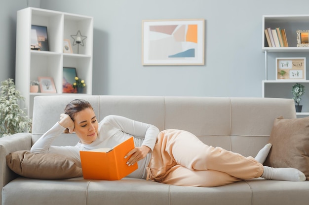 Mujer hermosa joven en ropa de casa relajándose en un sofá en el interior de casa leyendo un libro feliz y positiva sonriendo alegremente pasando el fin de semana en casa