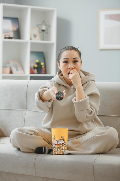 Mujer hermosa joven en ropa de casa sentada en un sofá en el interior de la casa con un cubo de palomitas de maíz sosteniendo a distancia viendo la televisión nerviosa y estresada mordiéndose las uñas