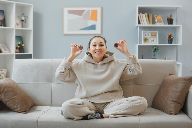 Mujer hermosa joven en ropa de casa sentada en un sofá en el interior de la casa sosteniendo galletas mirando a la cámara feliz y positiva sonriendo ampliamente