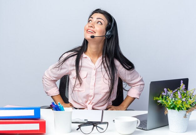 Mujer hermosa joven en ropa casual con auriculares y micrófono mirando hacia arriba sonriendo feliz y positivo sentado en la mesa con el portátil sobre la pared blanca trabajando en la oficina