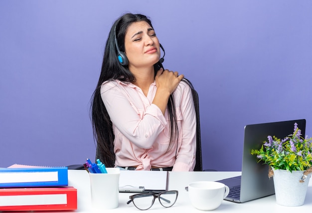 Mujer hermosa joven en ropa casual con auriculares y micrófono mirando malestar tocando el hombro sintiendo dolor sentado en la mesa con la computadora portátil sobre la pared azul trabajando en la oficina