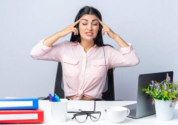 Mujer hermosa joven en ropa casual con auriculares y micrófono mirando molesto señalando con los dedos en sus sienes sentado en la mesa con la computadora portátil sobre la pared blanca trabajando en la oficina