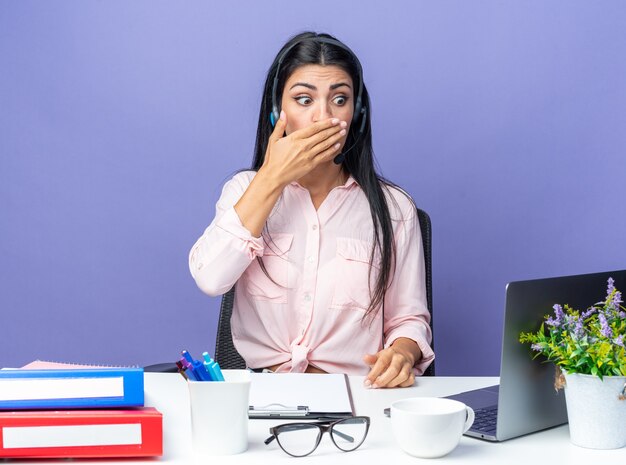 Mujer hermosa joven en ropa casual con auriculares con micrófono mirando la pantalla de la computadora portátil sorprendida cubriendo la boca con la mano sentada en la pared azul trabajando en la oficina