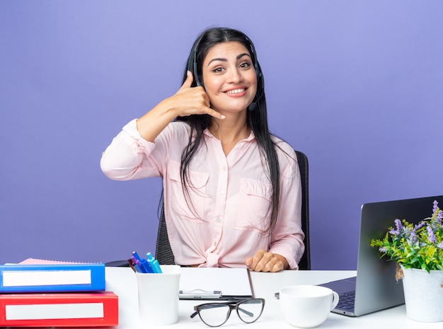 Mujer hermosa joven en ropa casual con auriculares con micrófono sonriendo haciendo gesto de llamarme sentado en la mesa con el portátil sobre la pared azul trabajando en la oficina