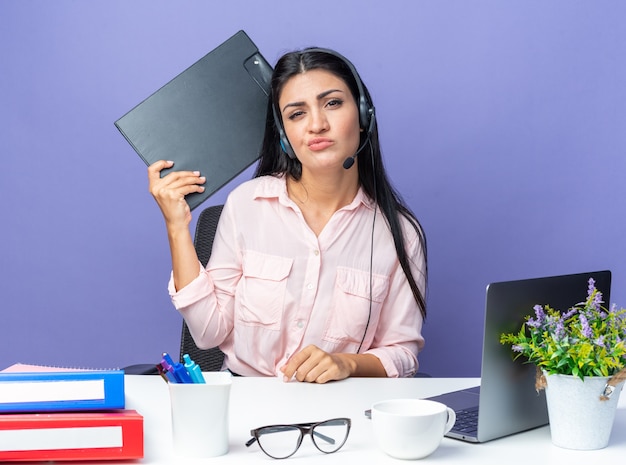 Mujer hermosa joven en ropa casual con auriculares y micrófono sosteniendo clipborad mirando confundido y disgustado sentado en la mesa con la computadora portátil sobre la pared azul trabajando en la oficina