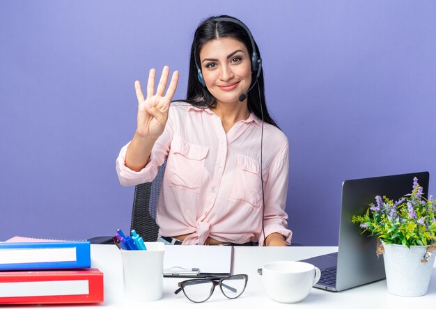 Mujer hermosa joven en ropa casual con auriculares mostrando el número cuatro sonriendo confiado sentado en la mesa con el portátil en azul