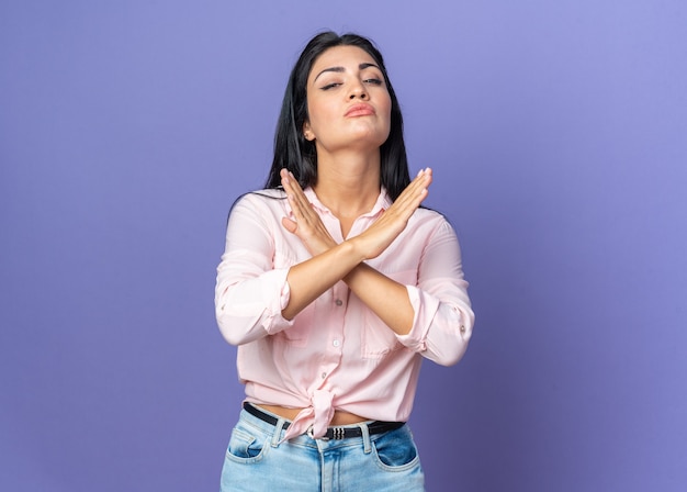 Mujer hermosa joven en ropa casual con cara seria haciendo gesto de parada cruzando las manos de pie en azul