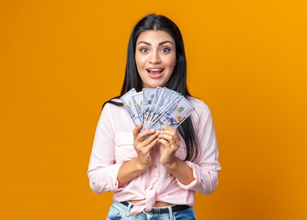 Mujer hermosa joven en ropa casual con dinero en efectivo feliz y positivo sonriendo alegremente de pie sobre la pared naranja